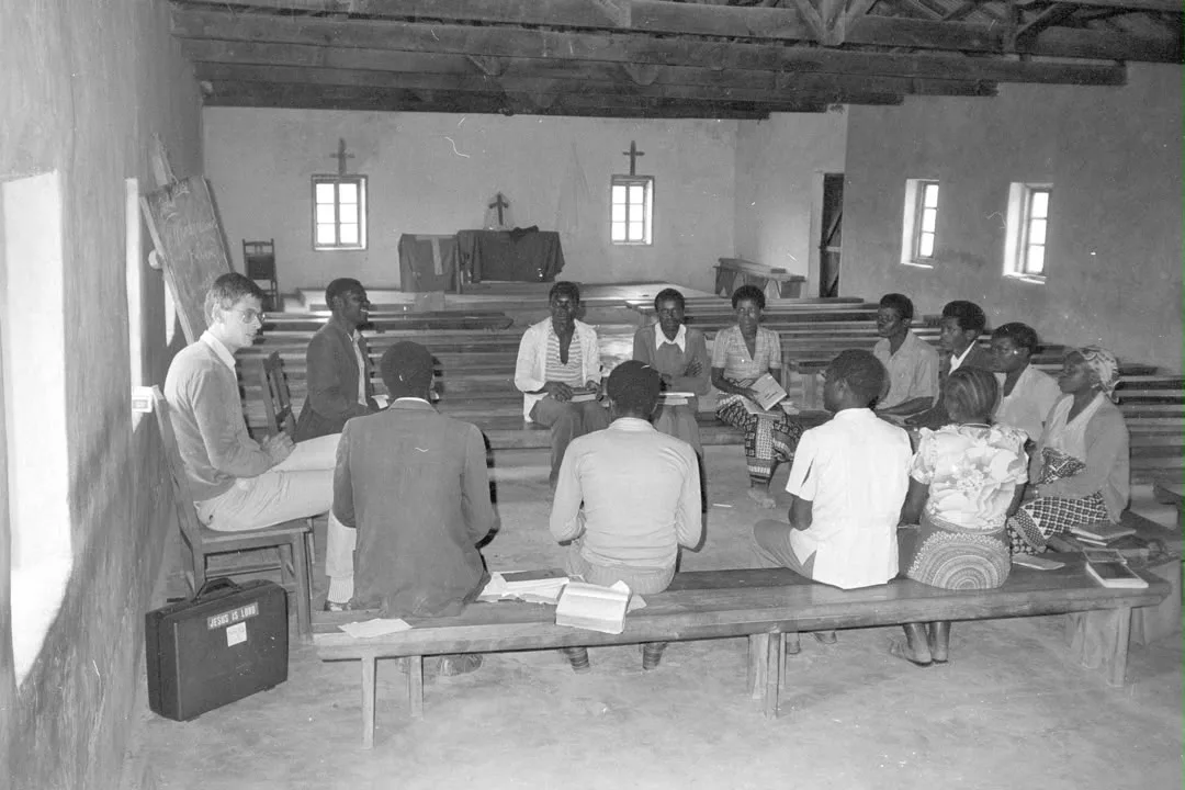 Picha ya zamani nyeusi-nyeupe: kikundi cha masomo kimeketi kwenye madawati ndani ya kanisa, mwalimu na ubao mbele